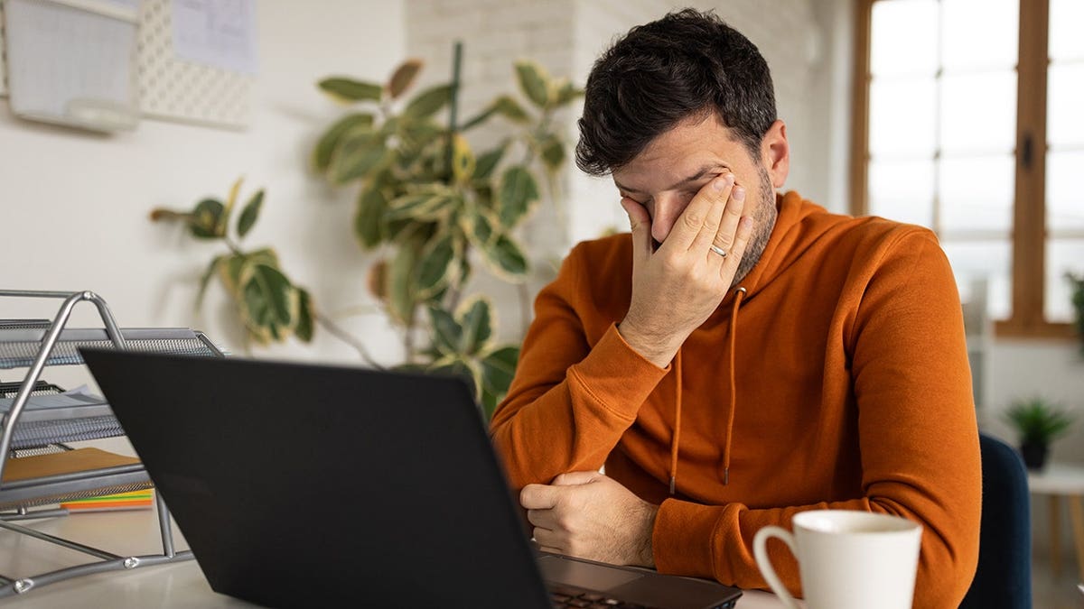 A mid adult Caucasian man showing fatigue sitting at a desk in his apartment office.