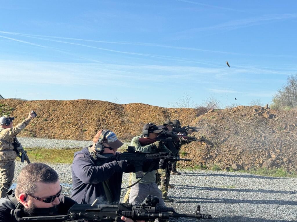 Line of shooters firing rifles at an outdoor range on a clear day, spent brass in the air.