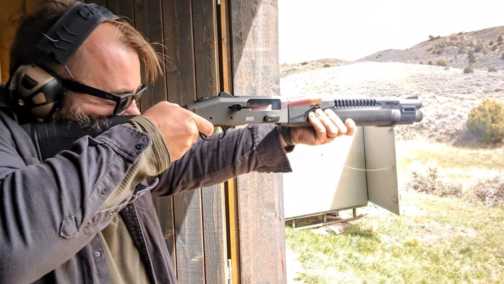 Person wearing earmuffs and glasses aiming a black firearm outdoors near a wooden structure.