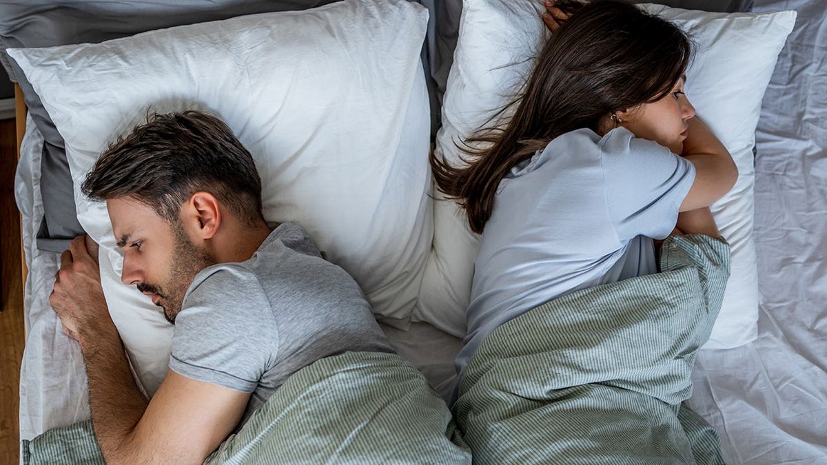 Young couple lies back to back in bed, appearing distant.