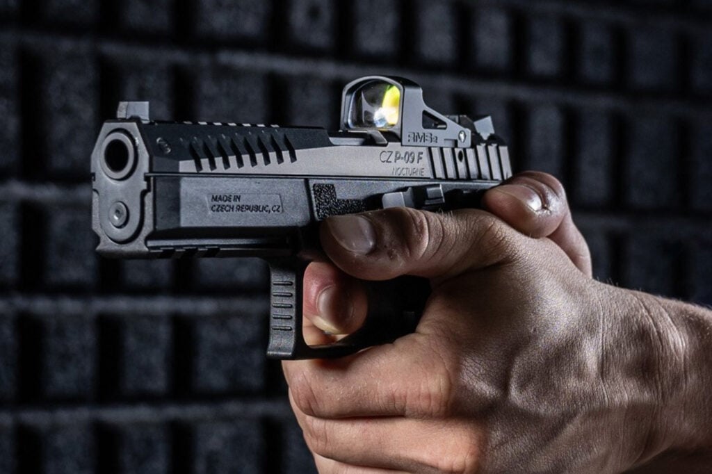Hand holding a black pistol with a mounted optic, illuminated light, and engraved text, against a dark brick background.