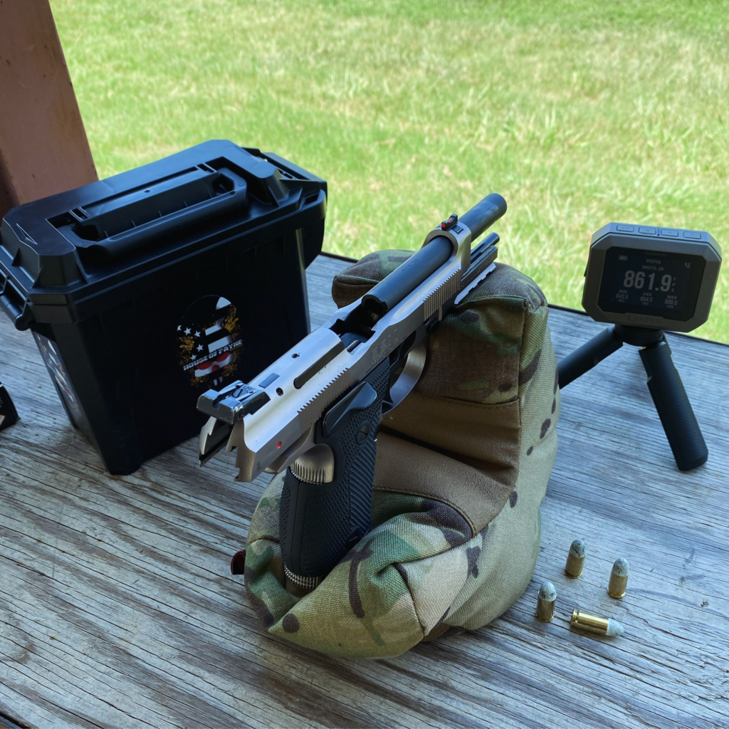 Stainless pistol slide in a camo rest beside a black case marked 'House of Fyre' and a chronograph display