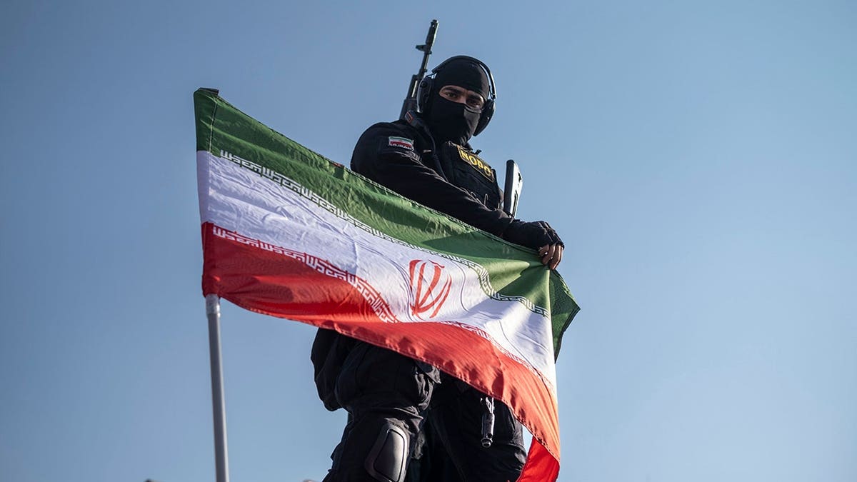 Armed Iran police special forces member adjusts flag on armored vehicle in Tehran.