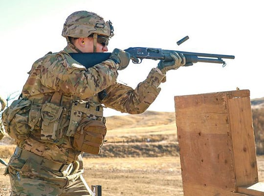 Soldier in camouflage aiming a blue shotgun at a wooden target in an outdoor setting.