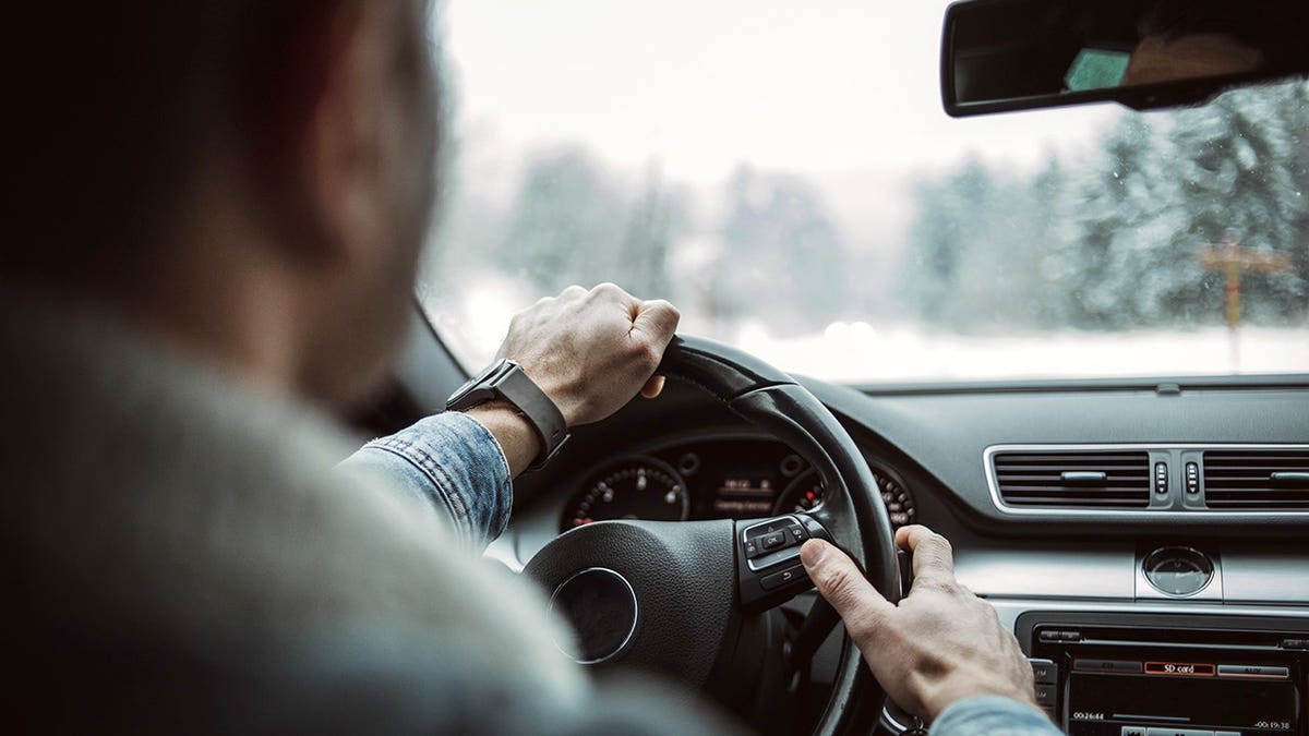 A man sits behind the wheel of a car in a snow-covered forest.