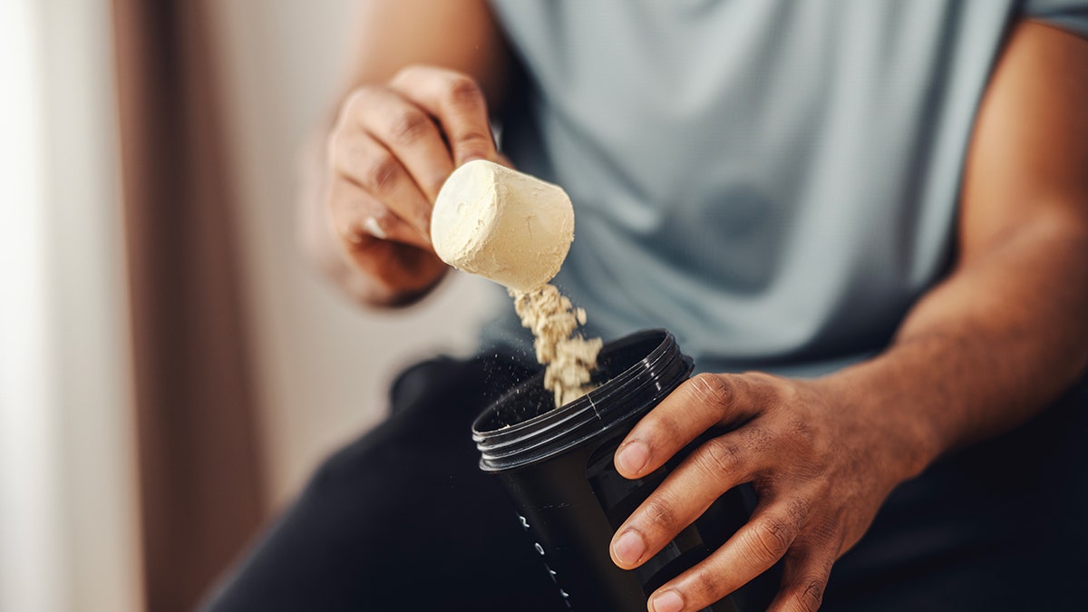 Adult man's hands scooping protein powder from a container into a black shaker bottle, preparing a nutritional supplement drink for workout recovery and muscle building