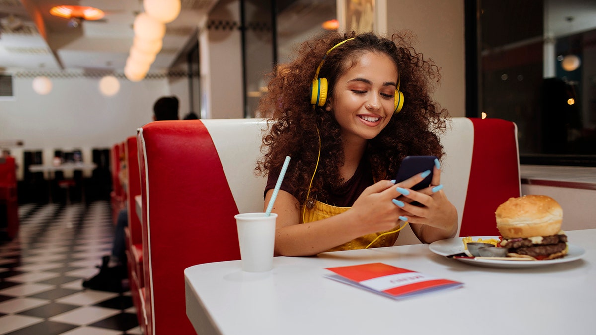 Young girl with headphones on smiling using her phone while dining alone at fast-casual restaurant with burger in front of her.