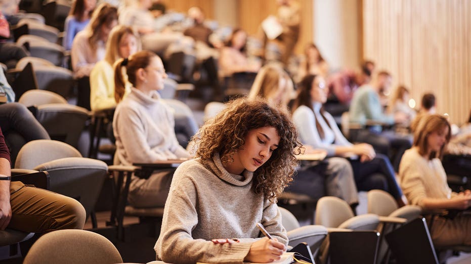College students sit in a lecture hall as a student takes notes during class.