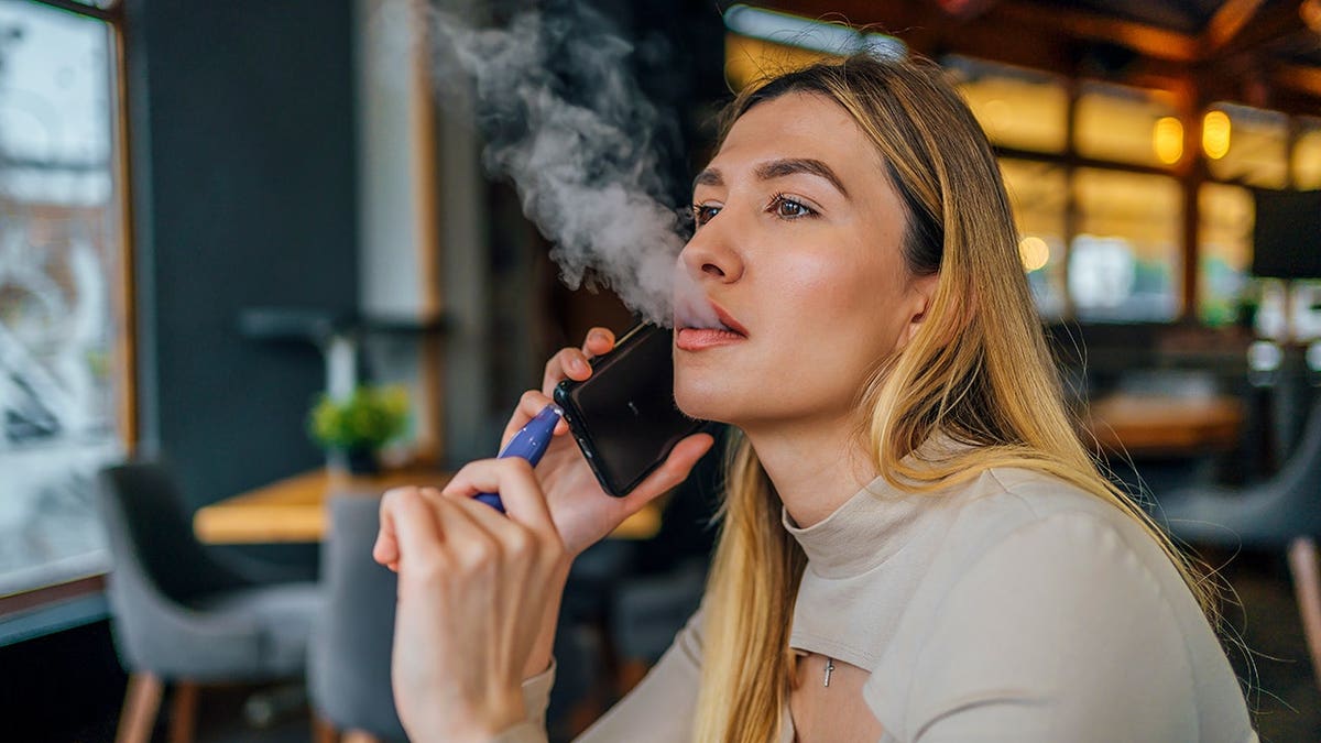 Woman smokes a disposable vape cigarette while talking on a smartphone.