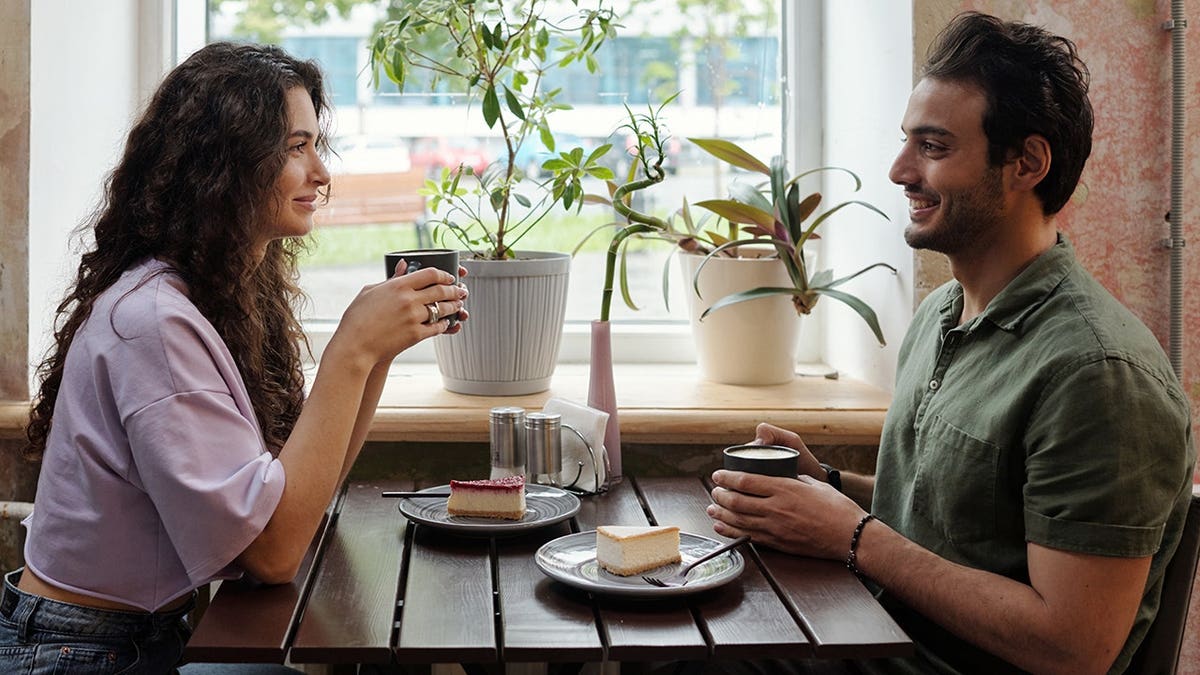 Young woman and smiling boyfriend sit at table with coffee cups, chatting.